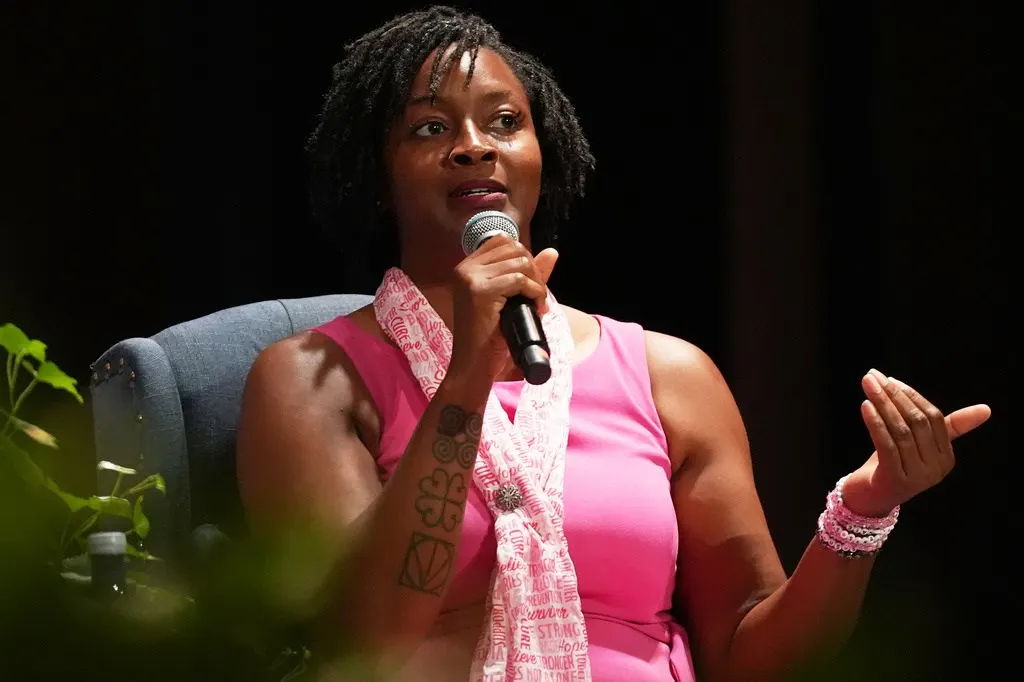A woman in a pink sleeveless top and scarf speaks into a microphone while gesturing, seated on stage.