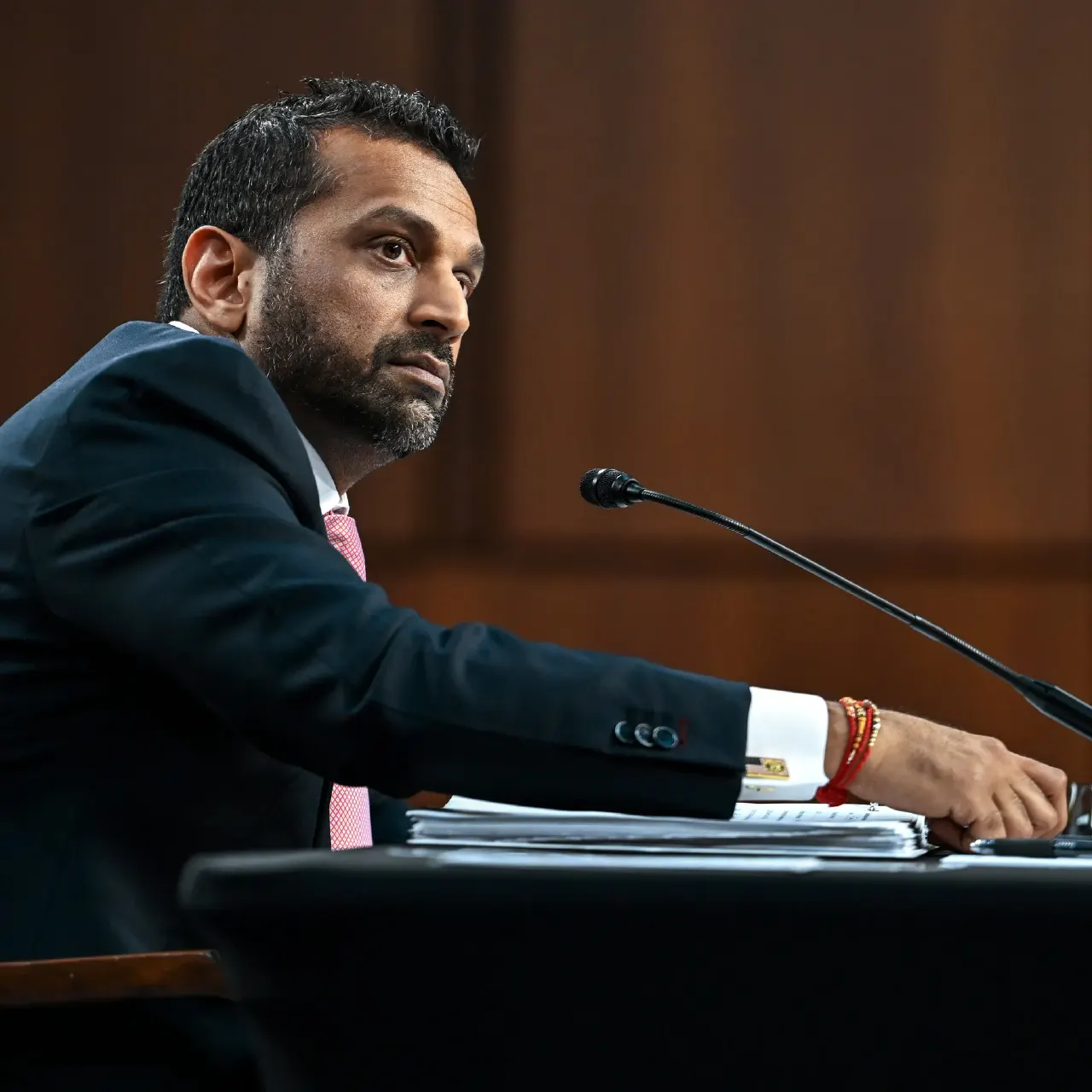 Man in dark suit and pink tie seated at a microphone, looking to the side.