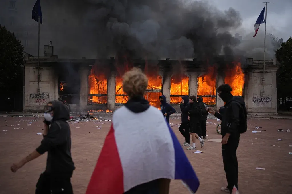 Masked protesters near a burning building, a person draped in a French tricolour flag, debris-strewn plaza.