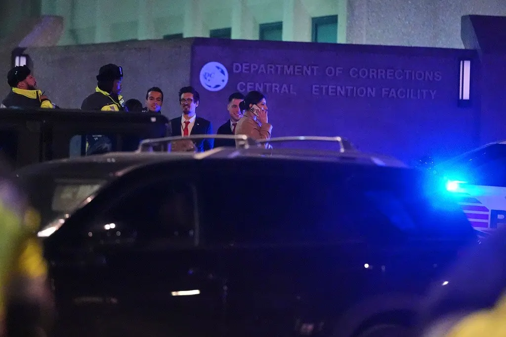 Men in suits and officers outside Central Detention Facility at night, with police lights and vehicles.