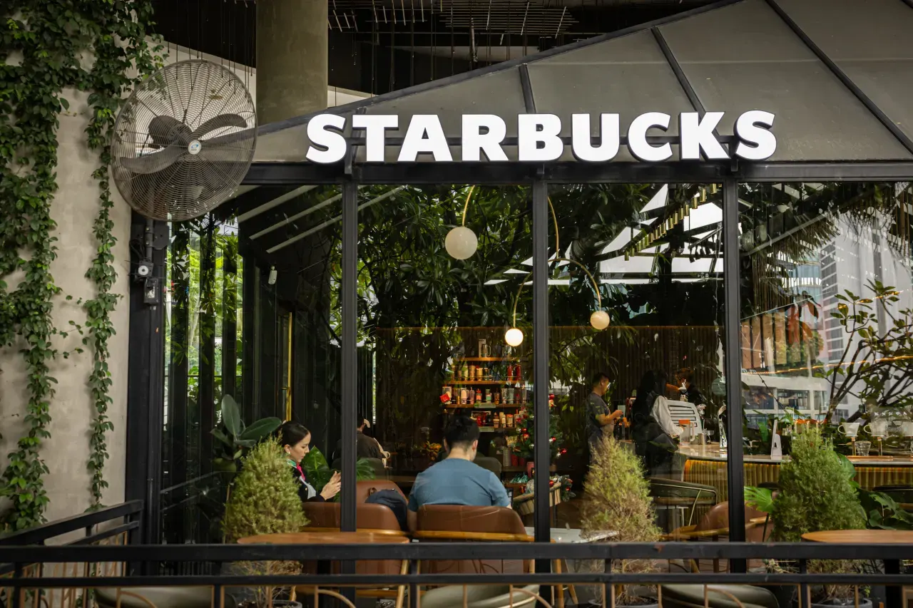 Starbucks cafe storefront with greenery, customers seated inside and a barista at the counter.