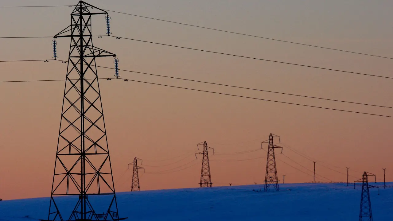 Silhouetted electricity pylons and power lines against a dusk sky over snowy hills.