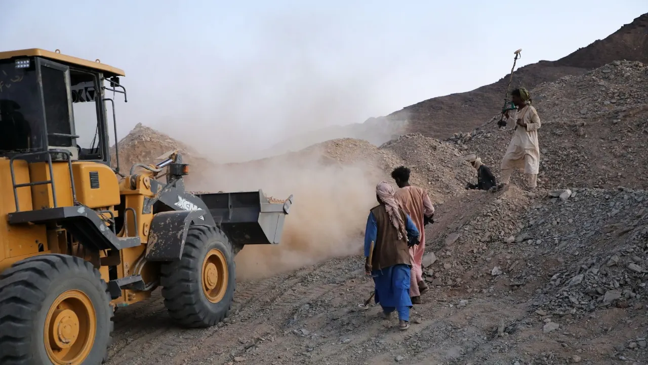Yellow loader scooping rubble at a dusty mining site while men in traditional dress walk nearby.
