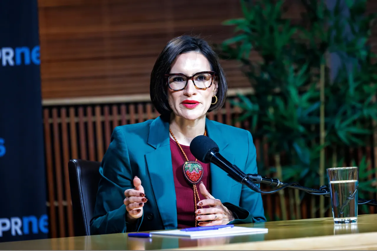 Woman in teal blazer speaks at microphone in studio, with PRNews backdrop and water glass.