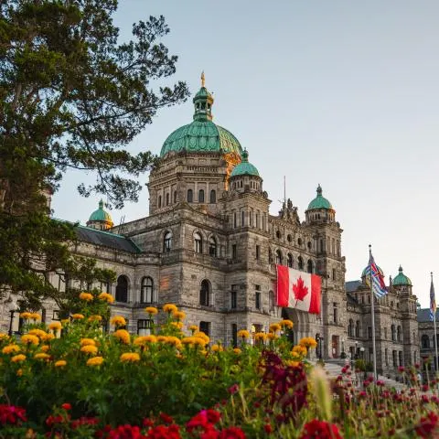 Stone legislative building with green domes, flowers, and Canadian and provincial flags at dusk.