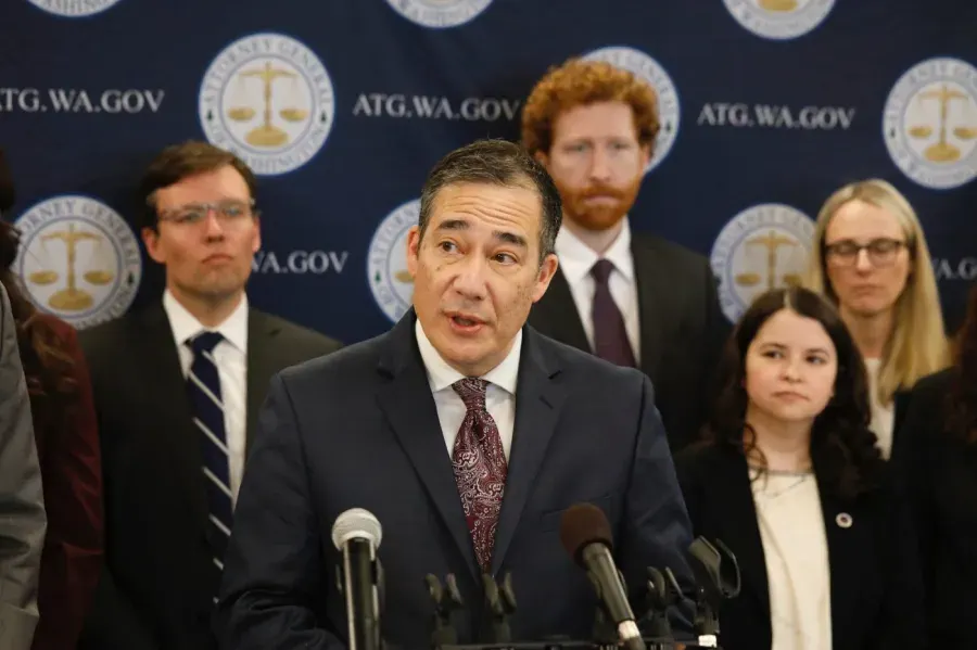 Man in suit speaking at podium before Washington Attorney General seal backdrop, with staff behind.