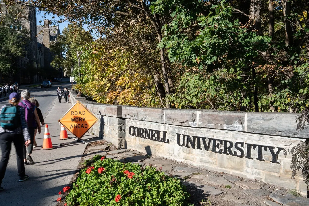 Stone wall with Cornell University lettering beside autumn trees, road work sign, and pedestrians on campus path.