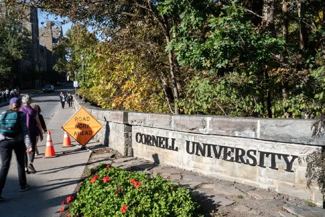 Stone wall with Cornell University lettering beside autumn trees, road work sign, and pedestrians on campus path.