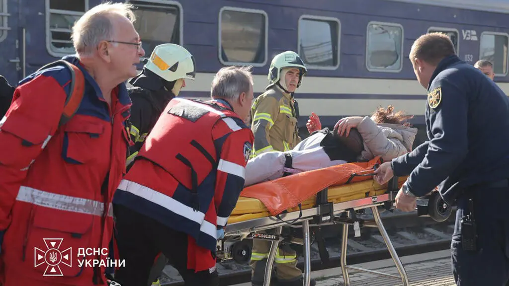 Rescue workers in red and firefighters move an injured person on a stretcher beside a train.