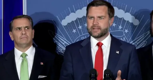 Man in navy suit speaks at podium before a blue seal backdrop, another man beside him.