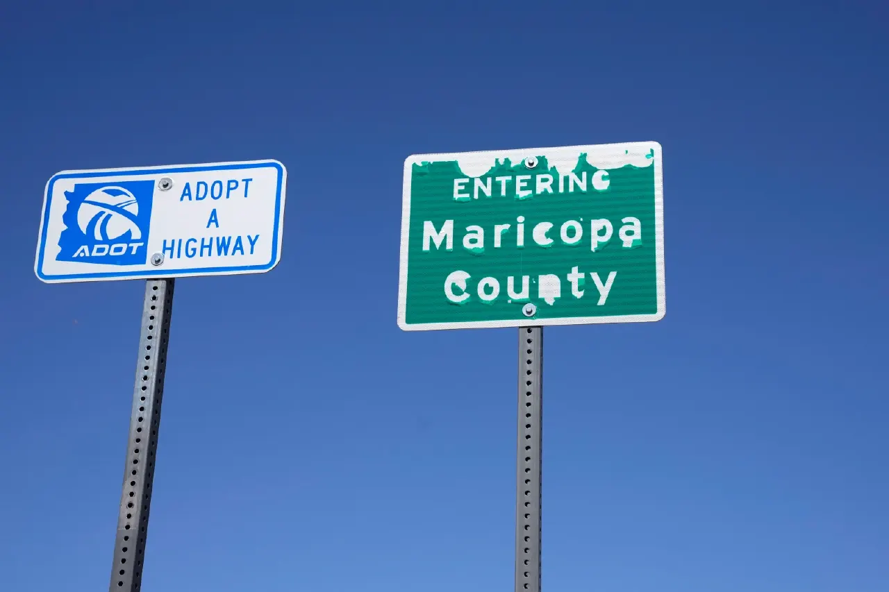 Blue sky behind two roadside signs: an ADOT Adopt a Highway sign and an Entering Maricopa County sign.