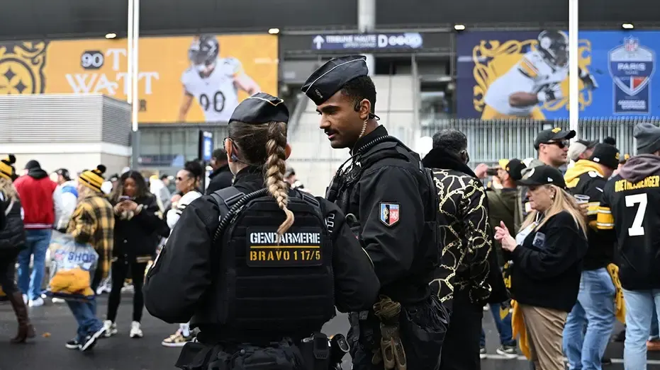 Gendarmes in tactical vests stand outside a stadium as Steelers fans walk past, with NFL Paris banners behind.