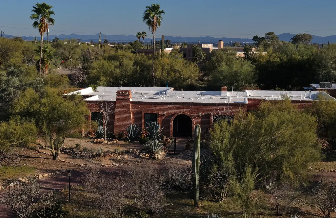 Aerial view of a red brick desert home with flat roof, surrounded by palms, cacti and mountains.