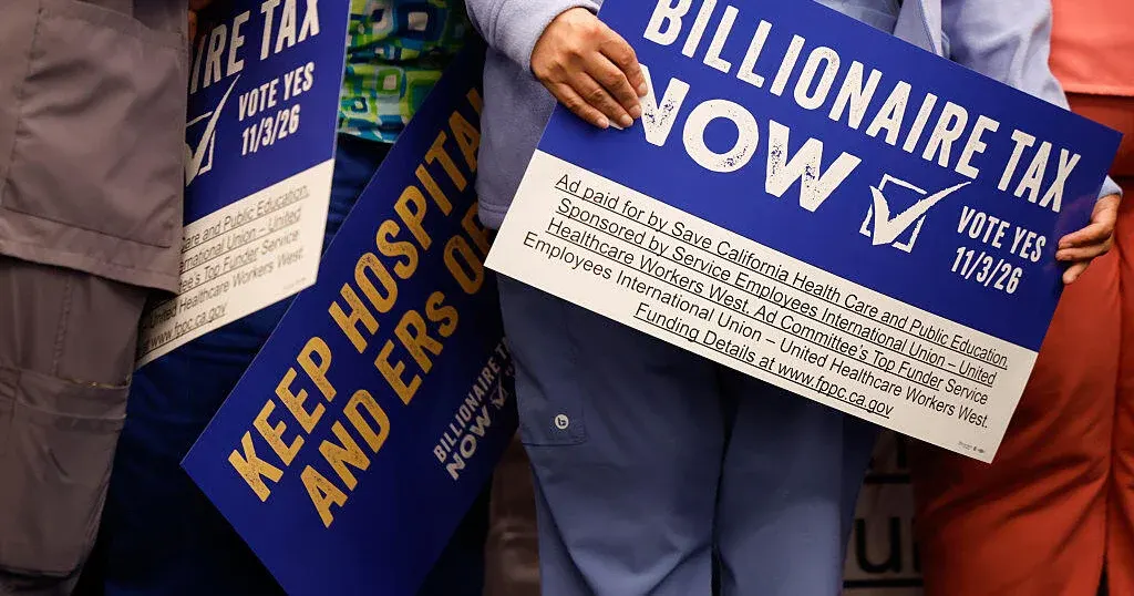 Crowd holds blue signs reading "Billionaire Tax Now" and "Keep Hospitals and ERs Open," faces cropped out.