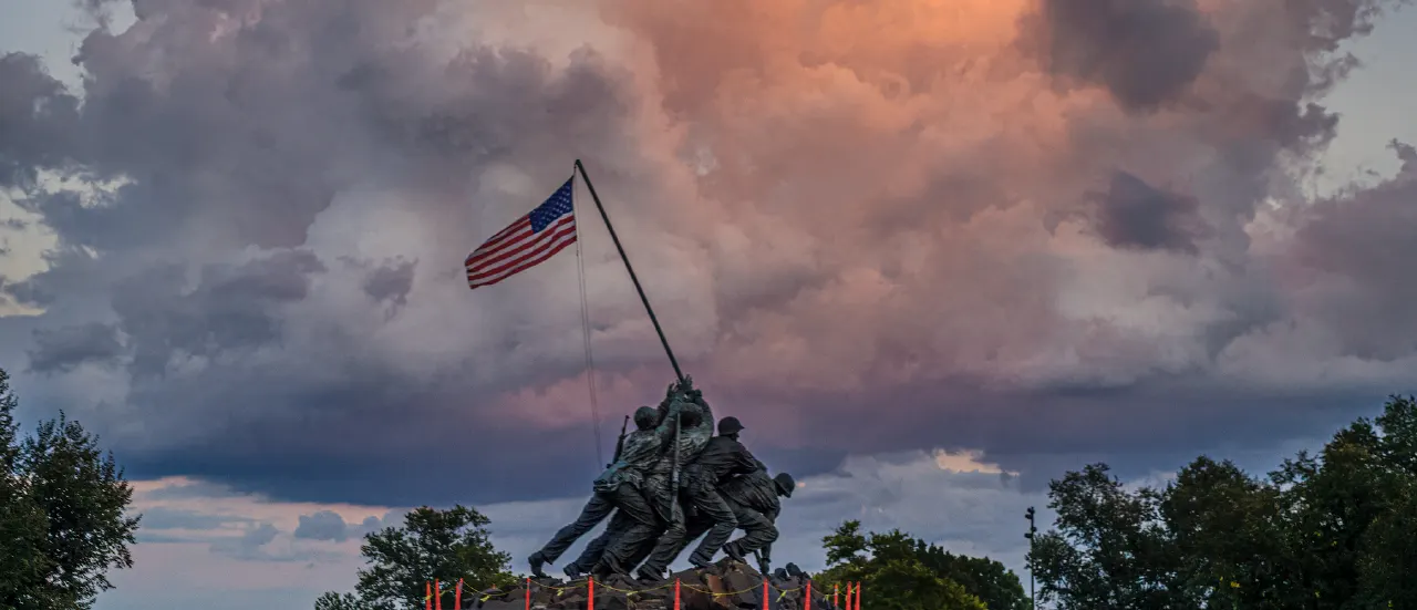 Bronze Iwo Jima memorial statue with American flag against dramatic sunset clouds and trees.