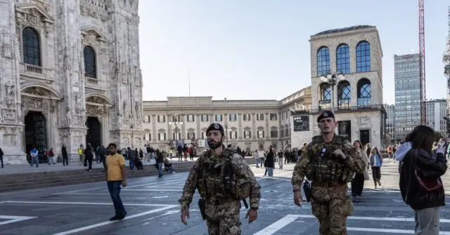 Two uniformed soldiers in camouflage walk through a Milan piazza past a cathedral and pedestrians.