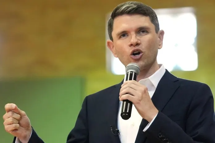 A man in a dark suit speaks into a handheld microphone, gesturing, against a green backdrop.