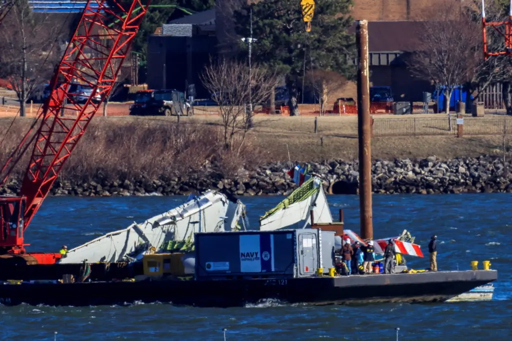 Aircraft wreckage on a barge in river being lifted by red crane, with a Navy-marked container and workers.