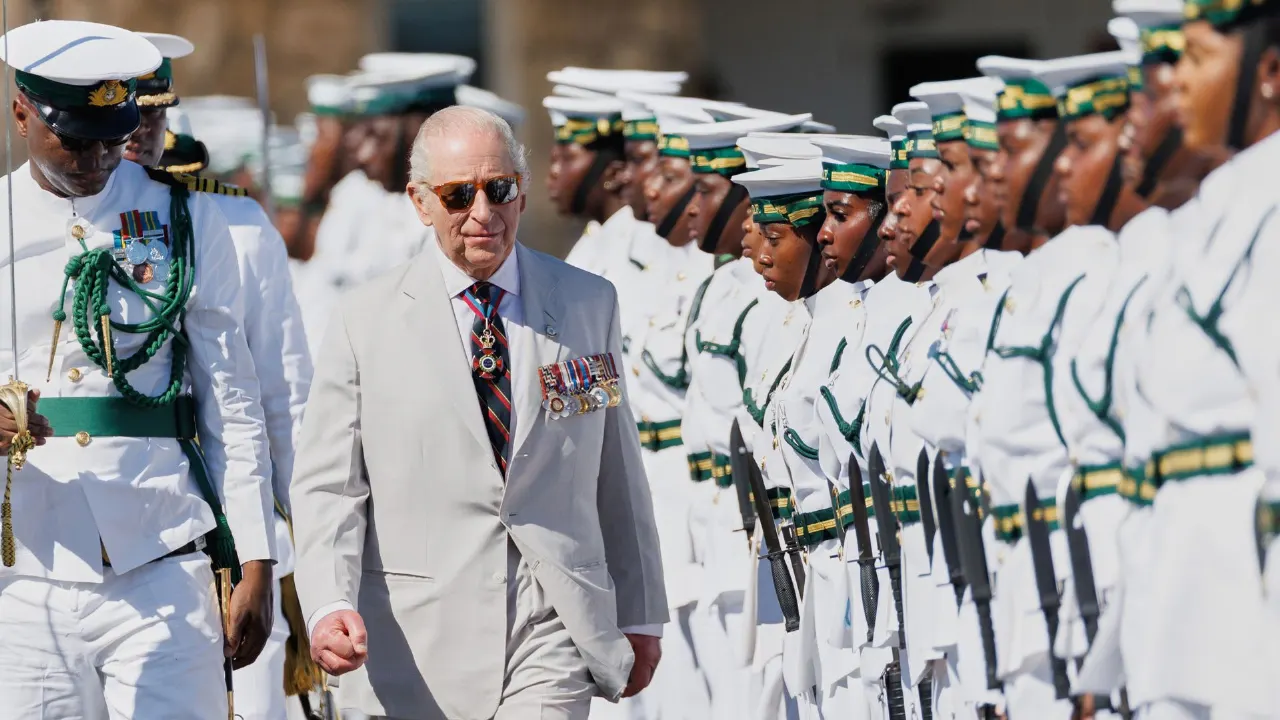 An older man in a light suit walks past a ceremonial honour guard in white uniforms with rifles.