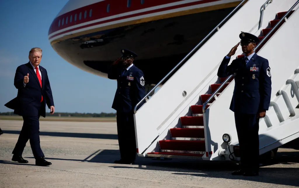 A man in a dark suit gives a thumbs up as uniformed officers salute beside an aircraft stairway.