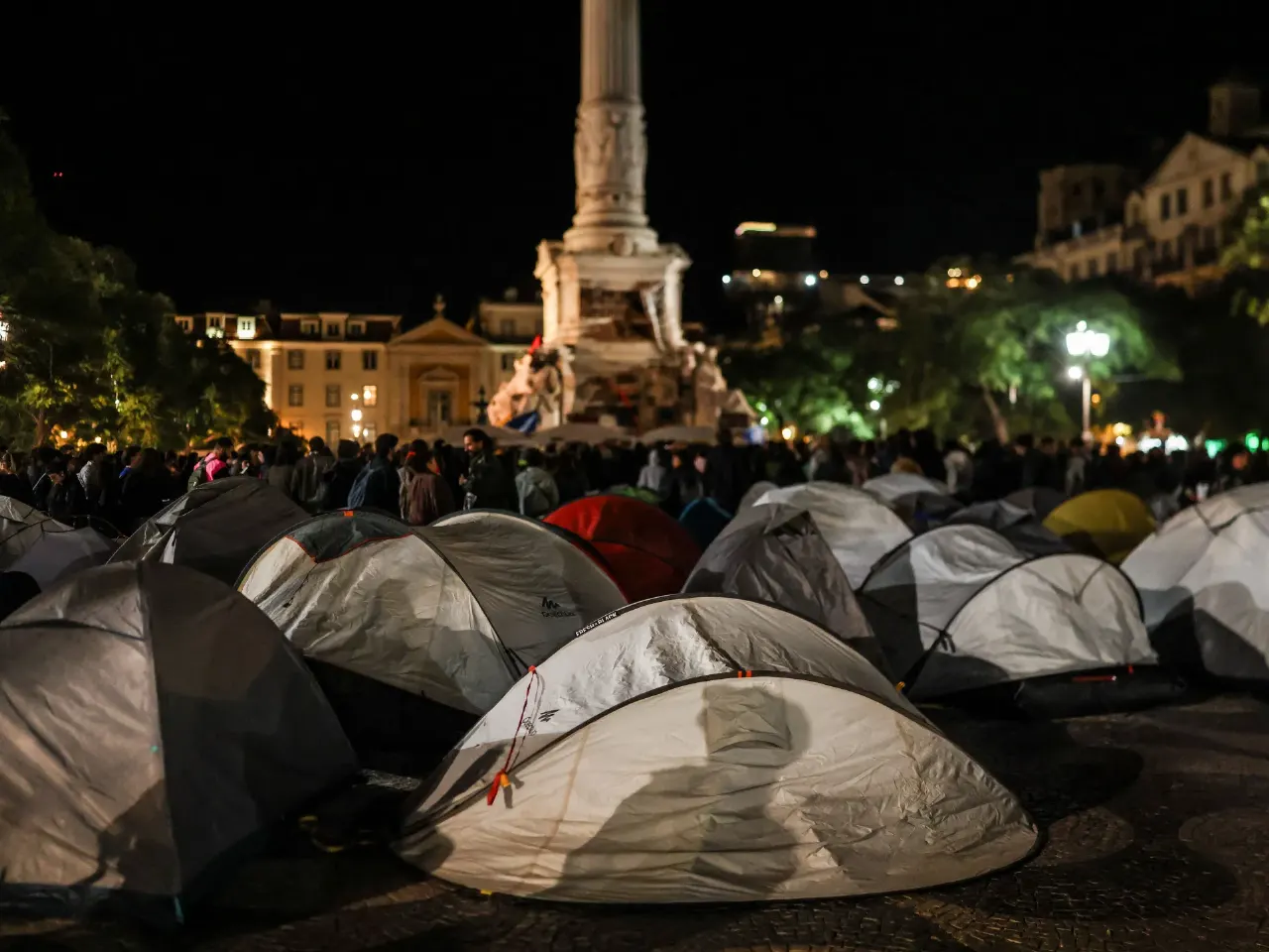 Night view of rows of tents in a city square, crowd gathered behind, monument column lit.