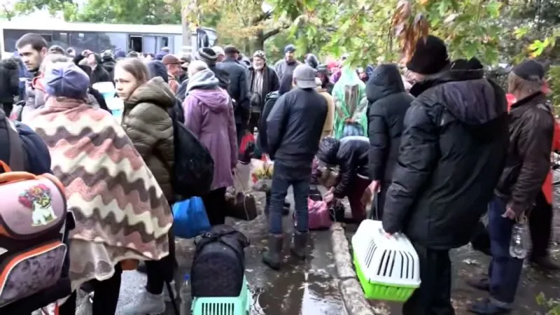 Crowd of evacuees with bags and pet carriers on a wet street beside a bus.