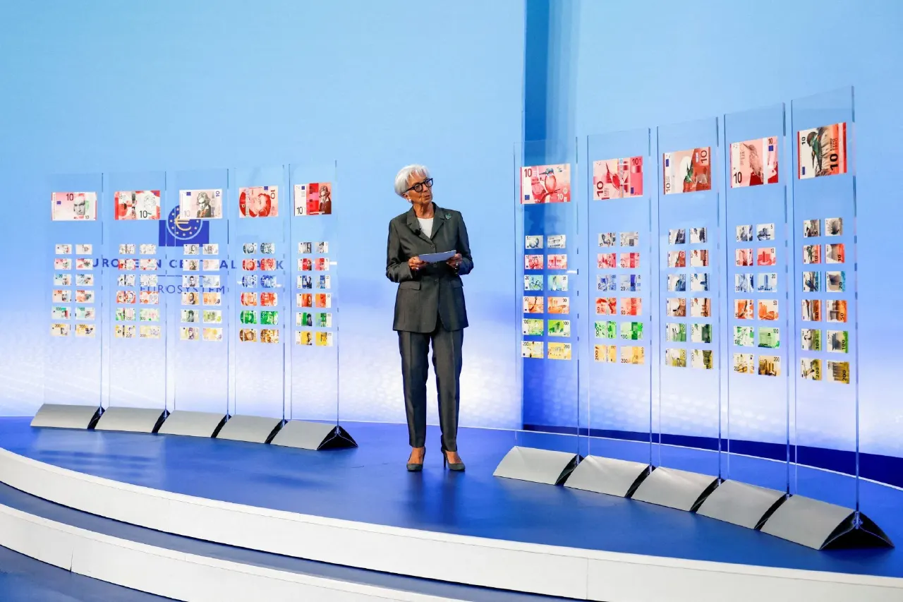 Woman in grey suit speaks on stage before displays of euro banknotes and ECB flag.