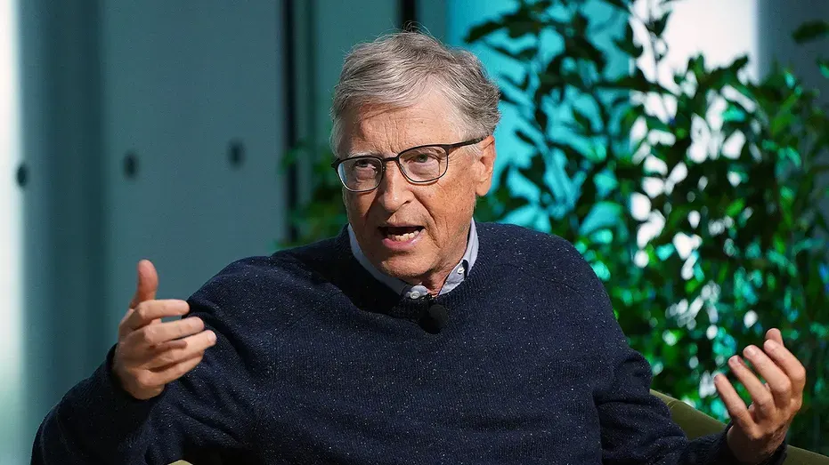 Older man with glasses in navy sweater gesturing mid-speech before green plants indoors.