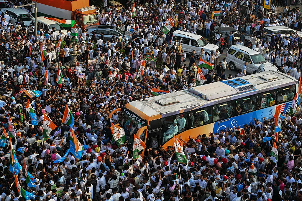 Large crowd waving tricolour flags surrounds a police bus in an aerial view of a street.