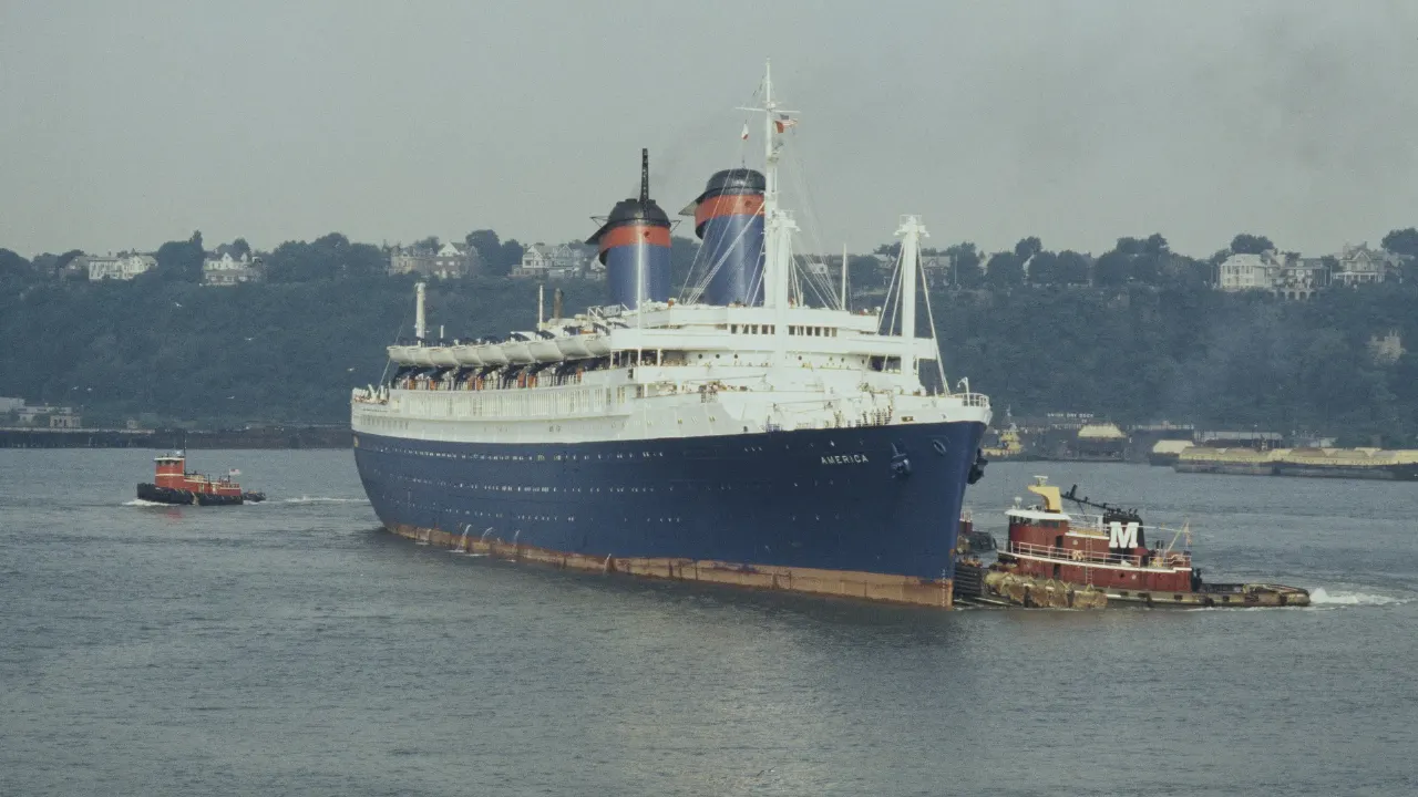 Large white and blue ocean liner with red funnels being towed by tugboats on a river.