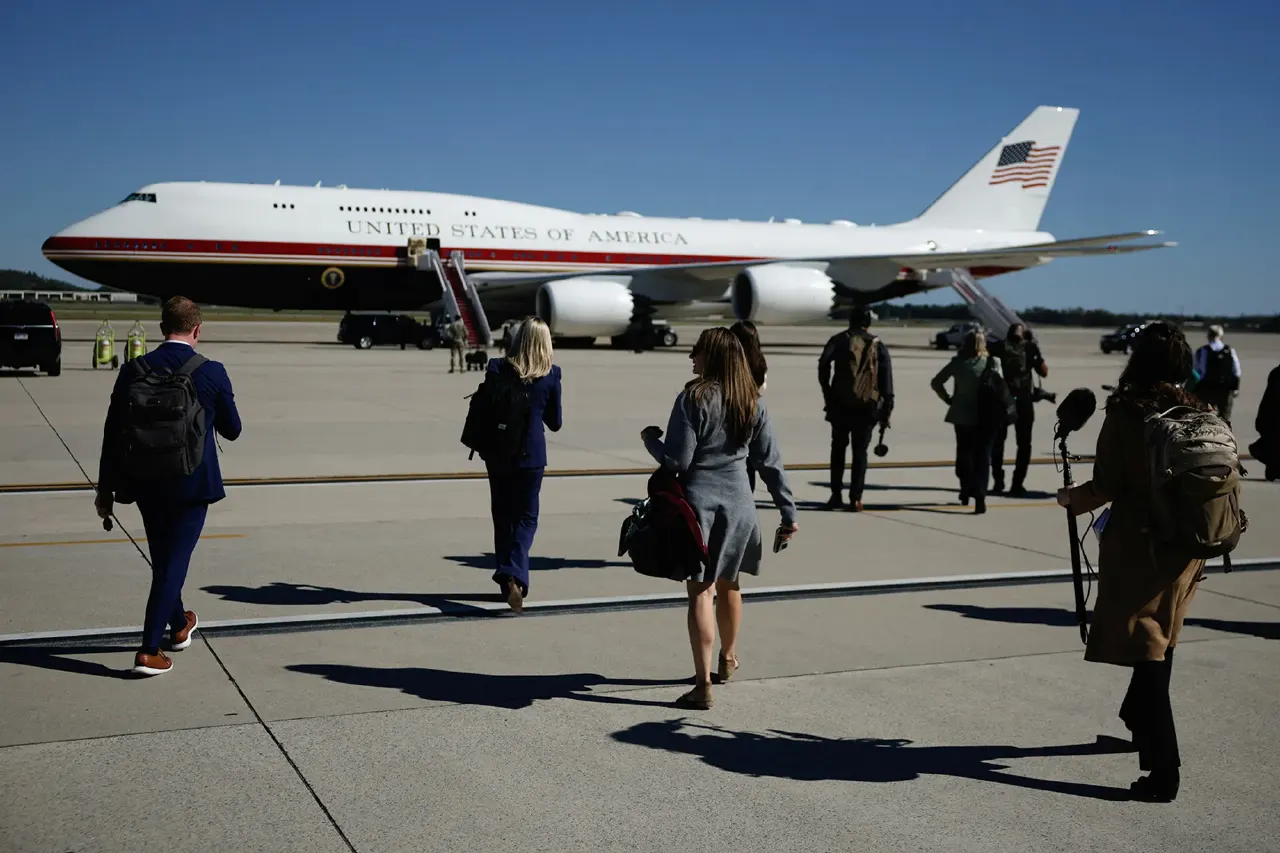 Reporters with bags walk across tarmac toward a US government aircraft bearing 'United States of America' livery.