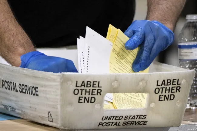 Gloved hands handling yellow envelopes in a United States Postal Service tray.