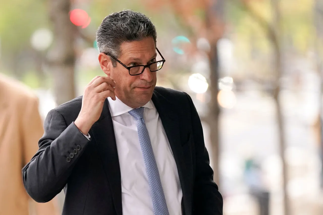 Man in dark suit, glasses and blue tie adjusting his hair on a city street.