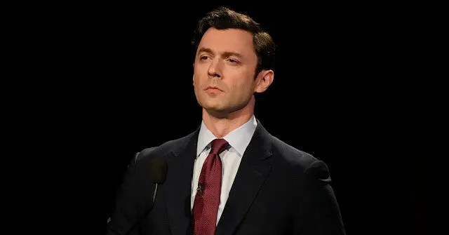 A man in dark suit and red tie speaking at a microphone against black background.