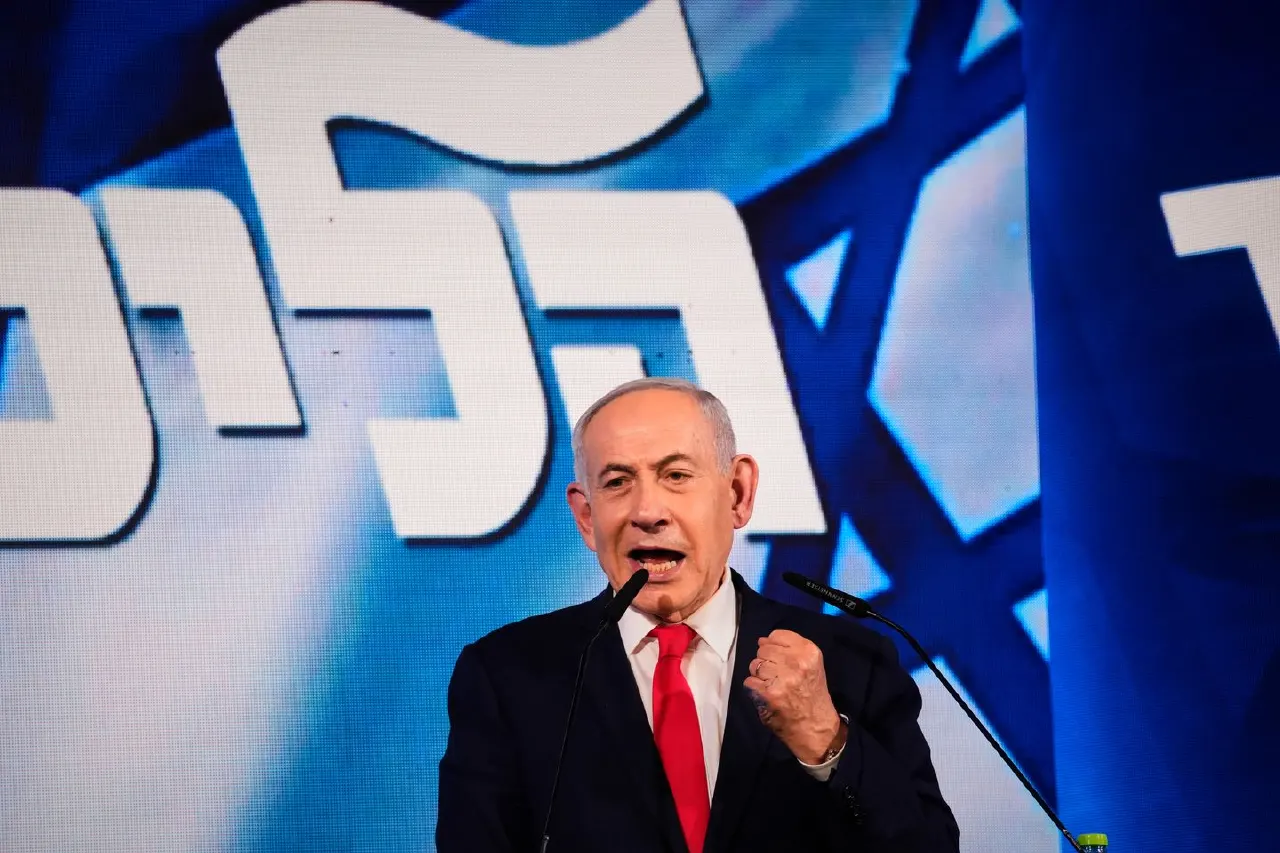 Man in dark suit and red tie speaking at podium before large blue party backdrop with Hebrew lettering