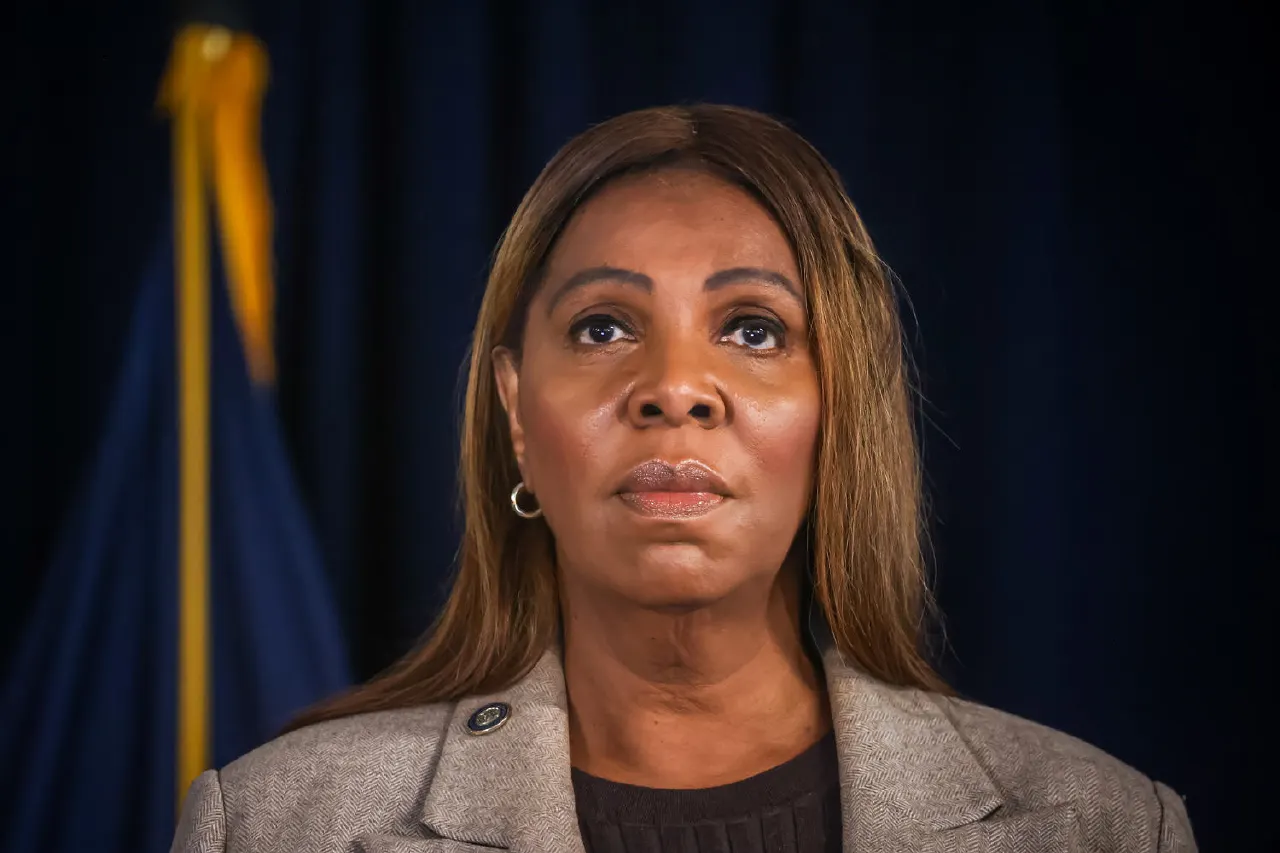 Woman in grey blazer with serious expression before dark blue backdrop and flag.