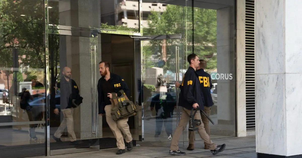 Several agents in FBI-labeled jackets exit a glass building entrance carrying bags and equipment.