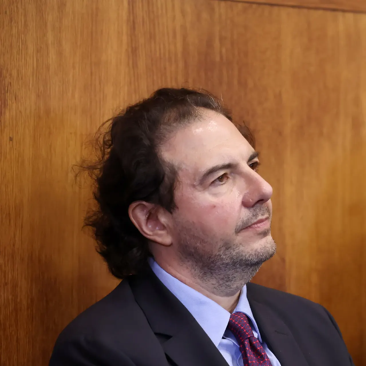 Man in dark suit and red tie seated before a wooden panel, looking to the side.