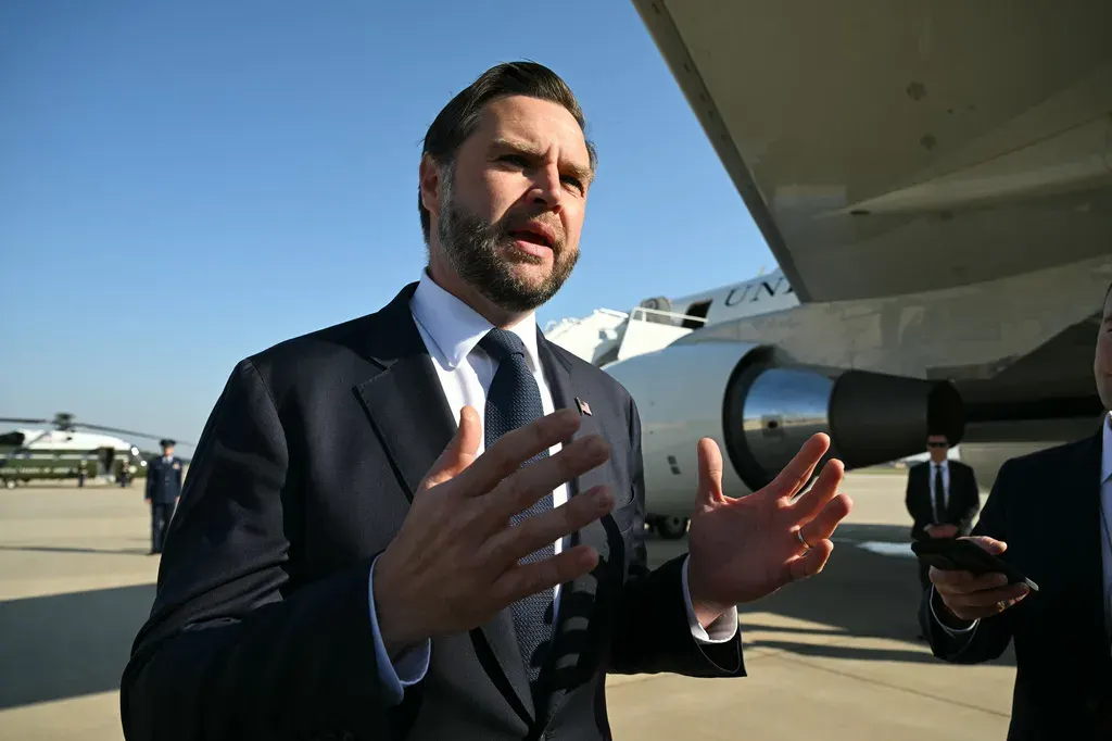 A bearded man in a dark suit gestures while speaking beside an aircraft on a tarmac.