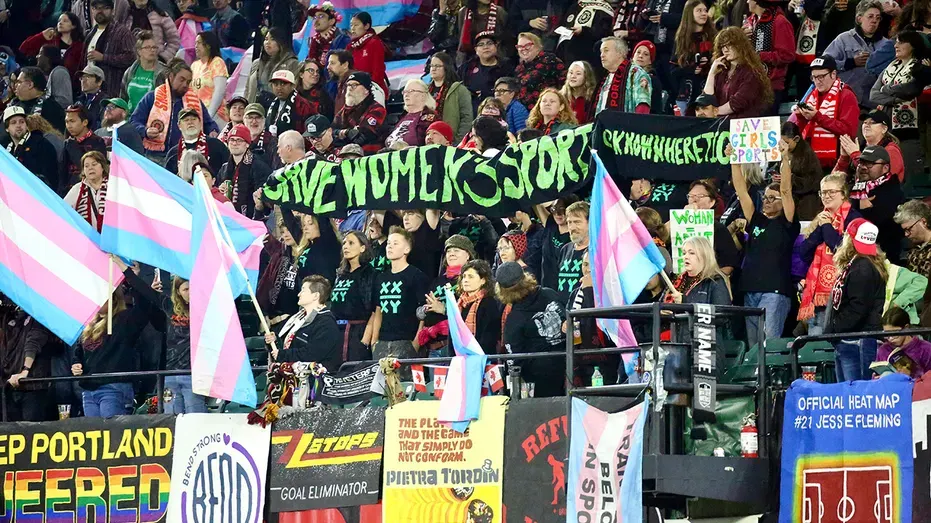 Stadium crowd holding a banner reading 'Save Women's Sports' beside transgender pride flags