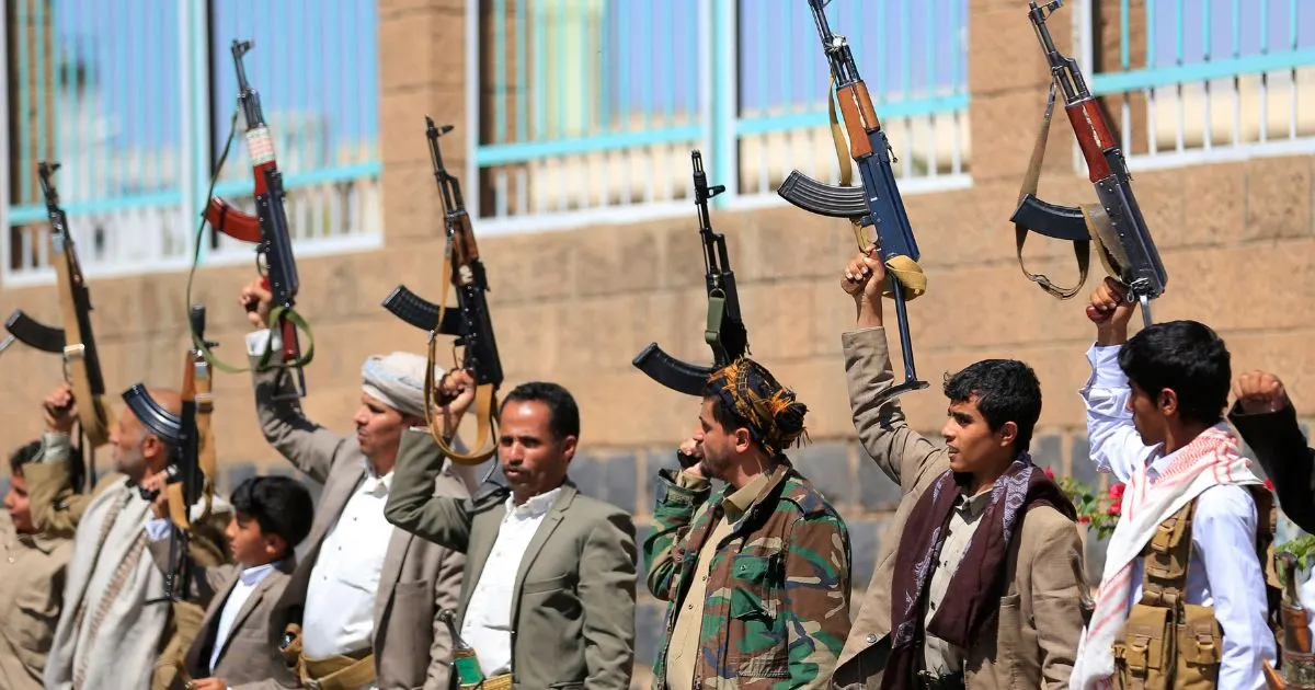 Armed men in civilian clothes raise assault rifles outdoors in front of a stone building.