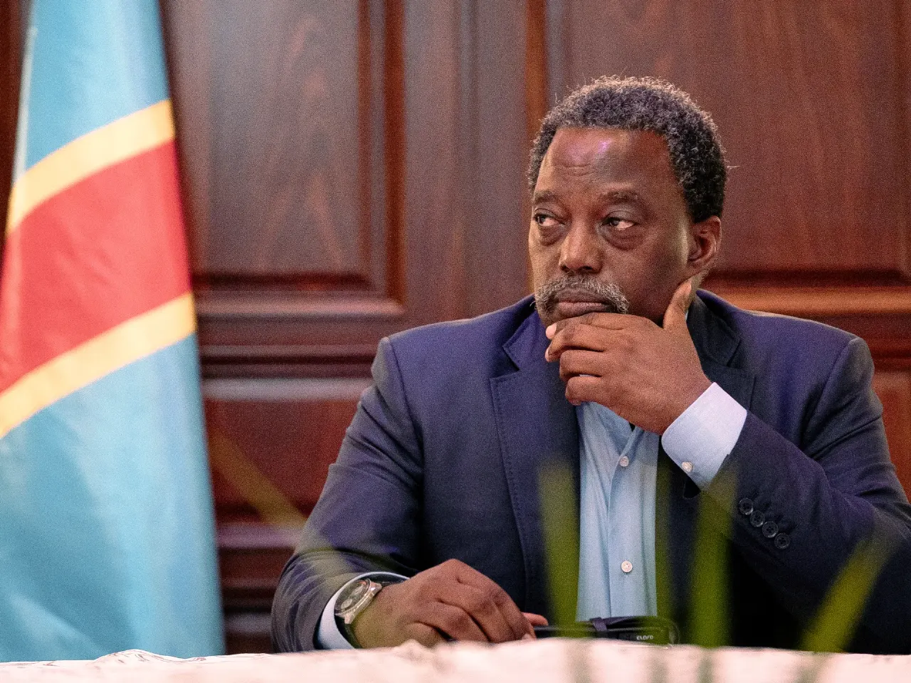 Man in dark suit, hand on chin, seated beside a blue, red and gold flag