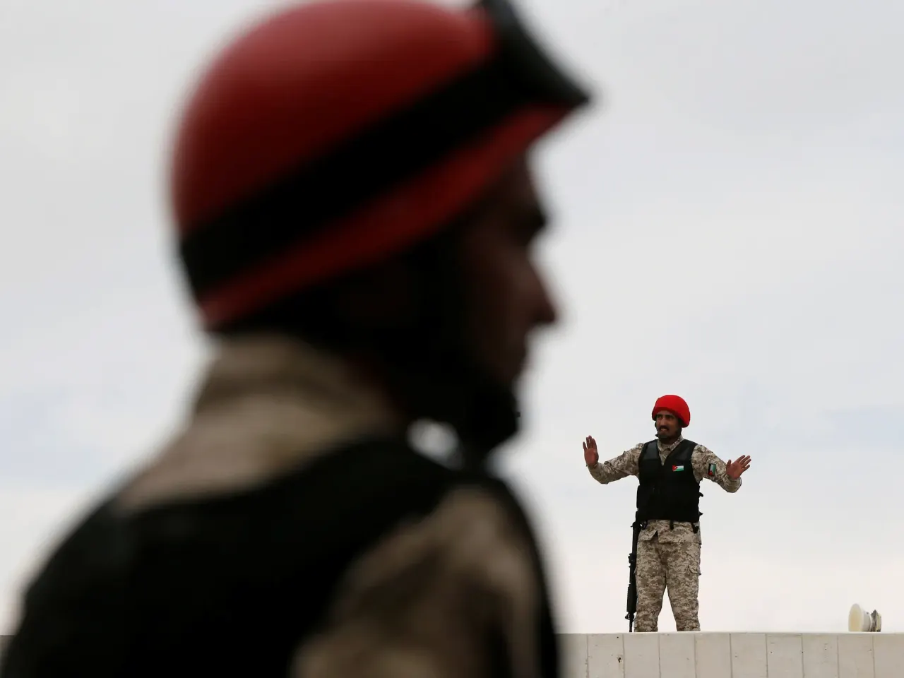 Blurred red-helmeted soldier in foreground; armed guard in camouflage gesturing on a wall above
