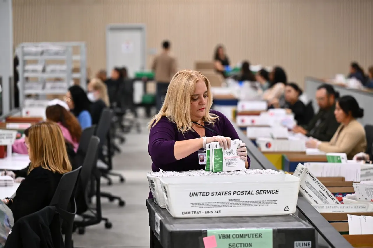 Election workers process mail ballots at tables, with a United States Postal Service tray in front.