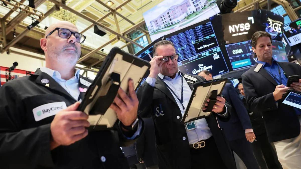 Traders on NYSE trading floor holding tablets, with NYSE and market screens behind them.