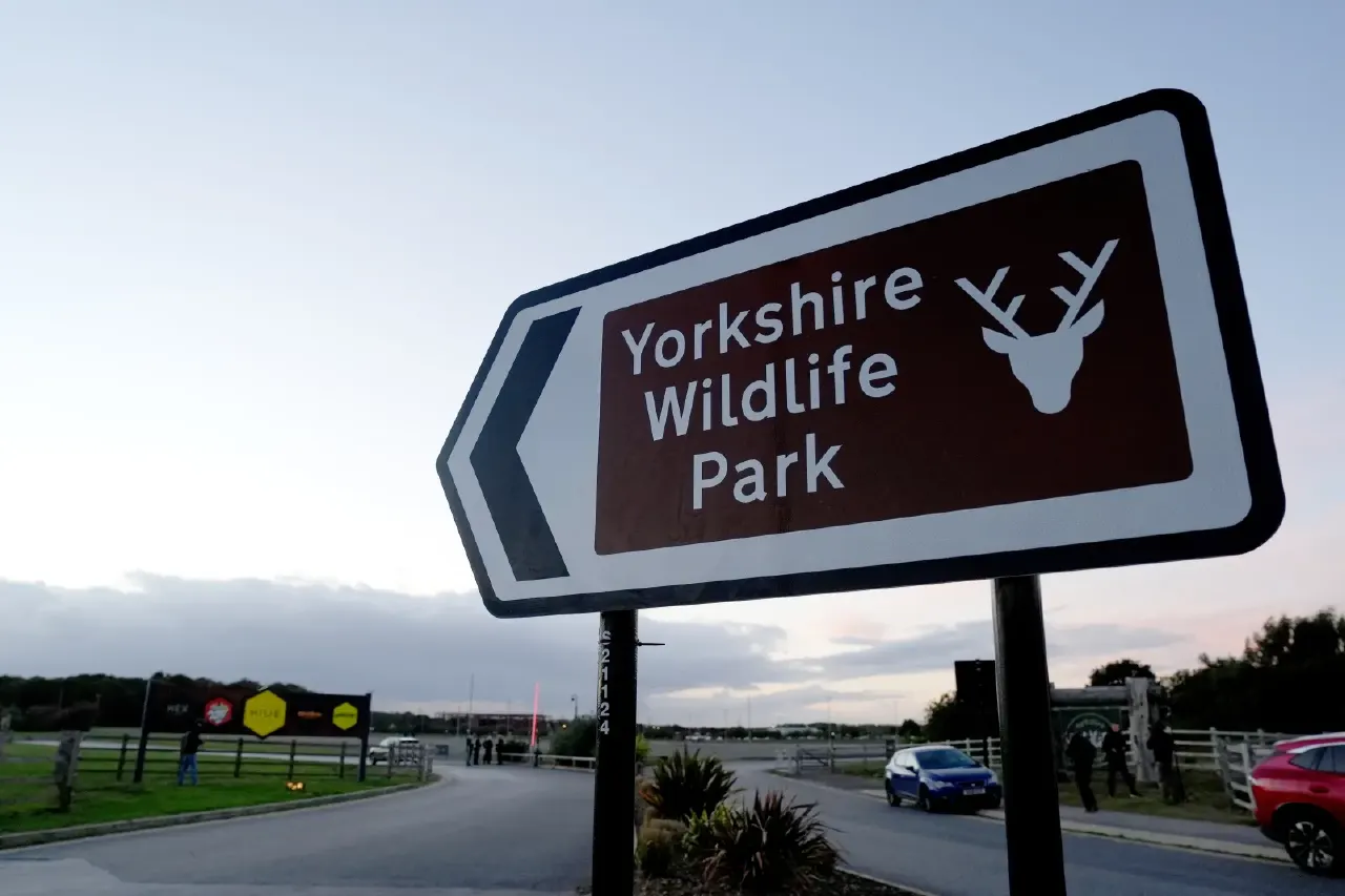 Directional sign for Yorkshire Wildlife Park at dusk, with parked cars and people nearby.