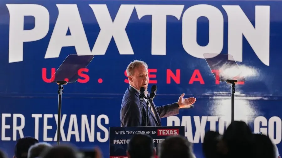 Man speaking at podium before large blue campaign banner reading PAXTON U.S. SENATE
