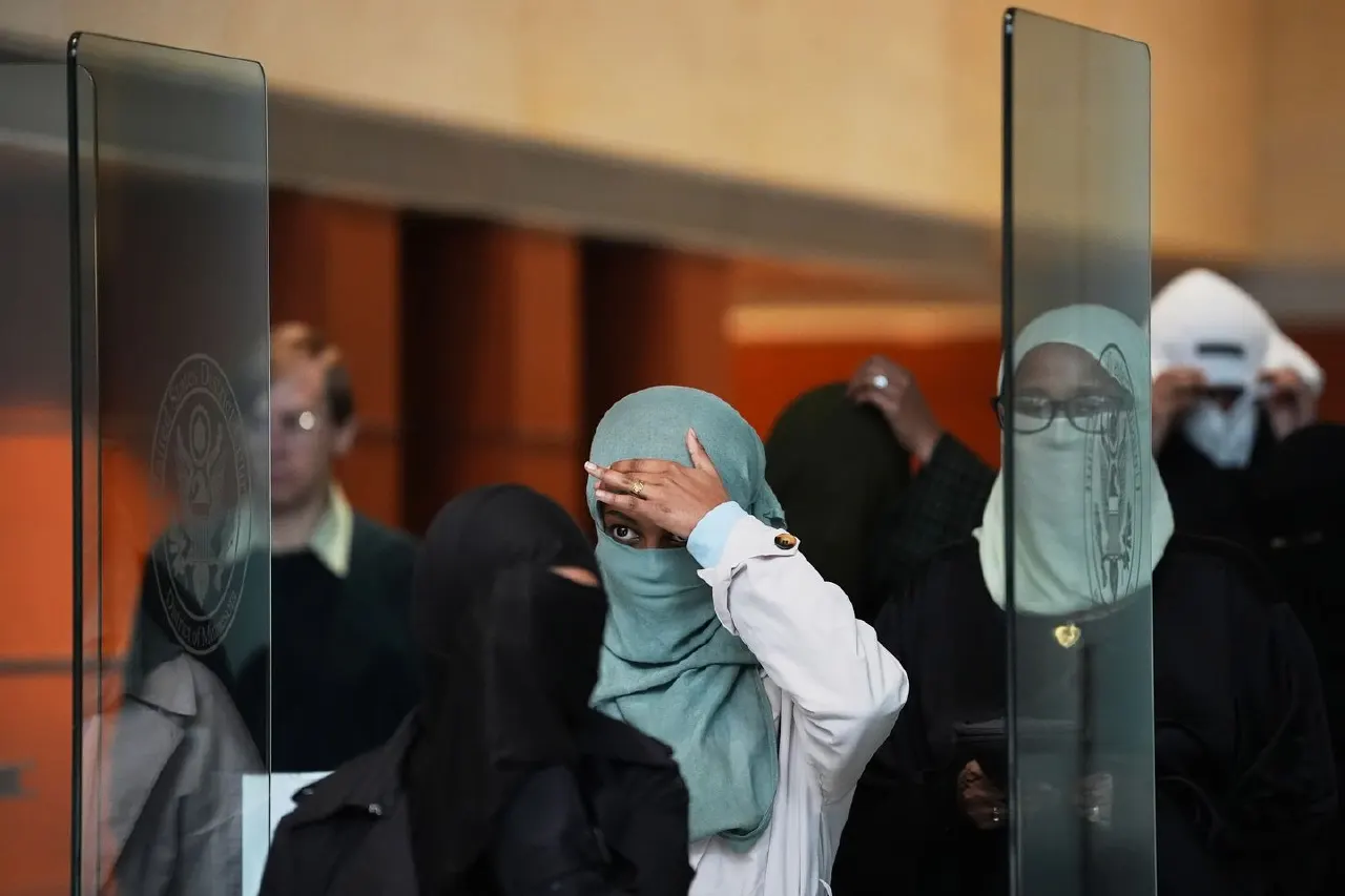 Several women in face coverings and headscarves stand behind glass partitions in a building lobby.