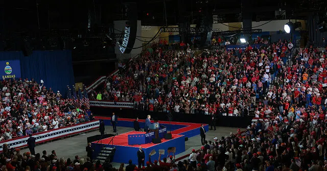 Large arena rally with a crowd in red shirts, a stage with a podium, and American flags.