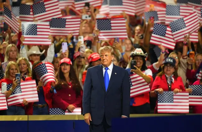 Political rally with crowd holding American flags behind speaker at podium.
